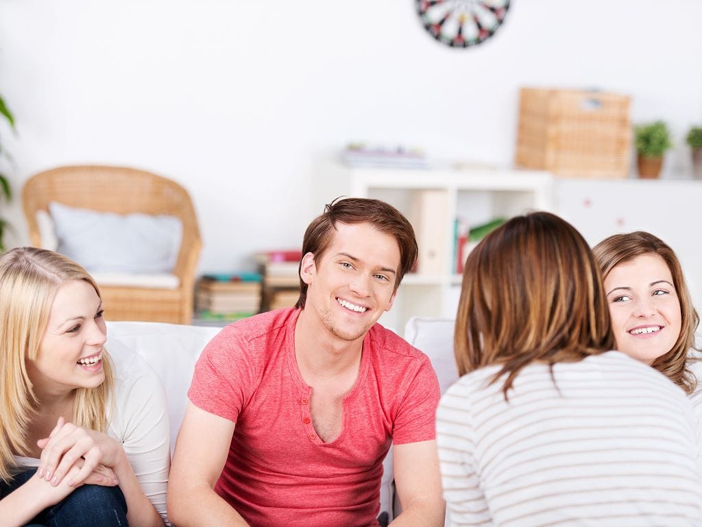 Junge Mitbewohnerinnen und Mitbewohner sitzen in einem hellen Wohnzimmer auf dem Sofa und unterhalten sich lachend